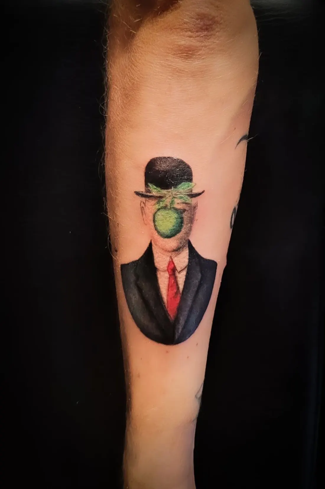Tatuaggio sull'avambraccio: rilettura a colori del «Figlio dell'uomo» di Magritte, l'uomo in bombetta con la mela verde sul volto.