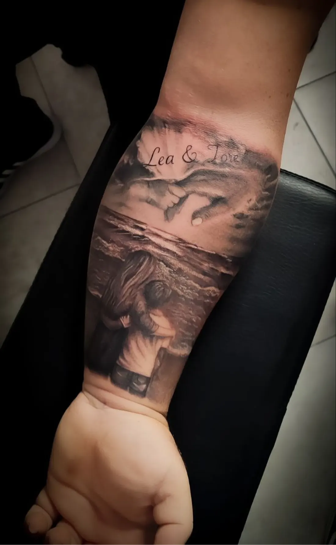 Tatuaggio sull'avambraccio: due figure abbracciate davanti al mare, mani che si sfiorano sopra l'orizzonte e la scritta «Lea & Tore».